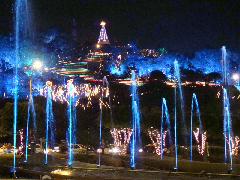 Colored Christmas light displays and fountains along the Medellín River at night, with a lit tree on the hillside